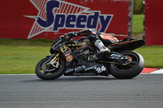 Eine Person in Helm und Schuhwerk fährt ein Motorrad auf einer von Gras umgebenen Rennstrecke, mit einer Tafel mit Text im Hintergrund.