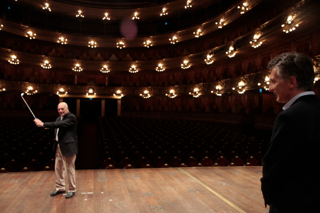 Ein älterer Mann in einem Anzug hält einen Golfschläger auf der Bühne, neben ihm steht ein weiterer älterer Mann, unter Deckenlampen mit vielen Sitzplätzen darunter.