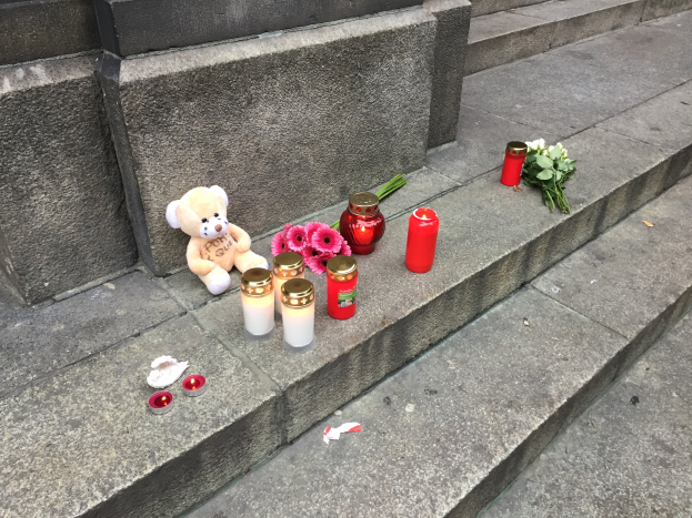 Ein Teddybär sitzt auf Treppenstufen neben angezönten Kerzen, Blumen und anderen Gegenständen und schafft eine friedliche Atmosphäre.