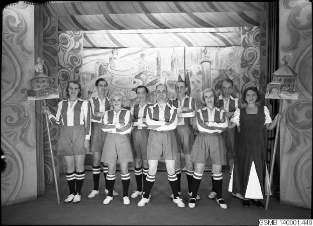 Eine Gruppe strahlender Frauen in einem 1940er-Soccer-Team posiert auf einer Bühne und hält Stöcke, mit einem Gemälde im Hintergrund, schwarz-weiß.