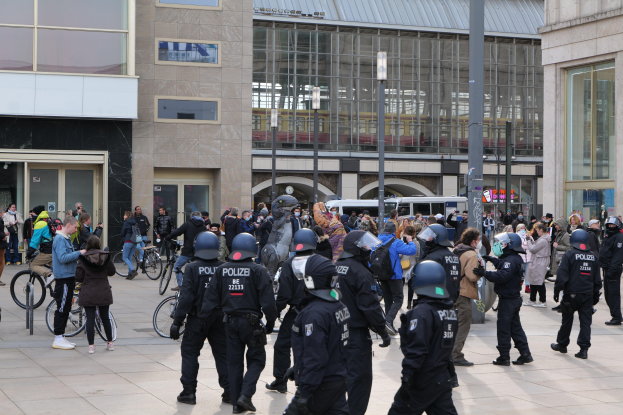 Eine Gruppe von Polizisten in Helmen steht vor einer Menge, die Fahrräder hölt, mit Glasfassade-Gebäuden, Laternenmästen und Fahrzeugen im Hintergrund.