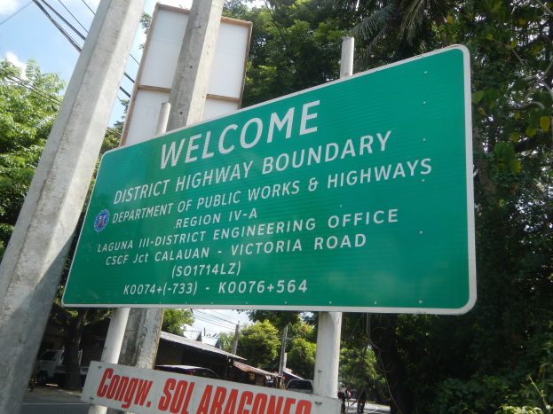 Ein grünes Schild mit der Aufschrift "Willkommen an der Kreisgrenze" ist an zwei weißen Pfählen befestigt, im Hintergrund sind Fahrzeuge, Gebäude und Bäume unter einem blauen und weißen Himmel zu sehen.
