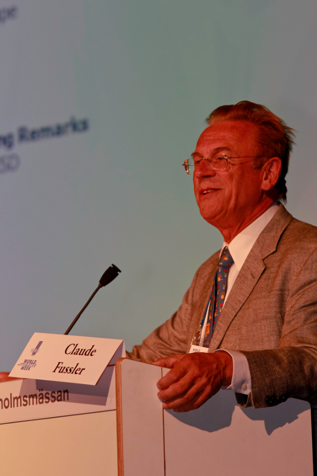 Ein Mann in einem weißen Hemd und einem grauen Blazer steht lächelnd vor einem Podium mit Mikrofon und Namensschildern, im Hintergrund ein Projektionsschirm oder Banner mit Text.