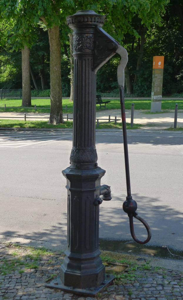 Eine altmodische Wasserpumpe in einem Park, umgeben von einer Straße, Gras, einer Bank, einem Brett und Bäumen.