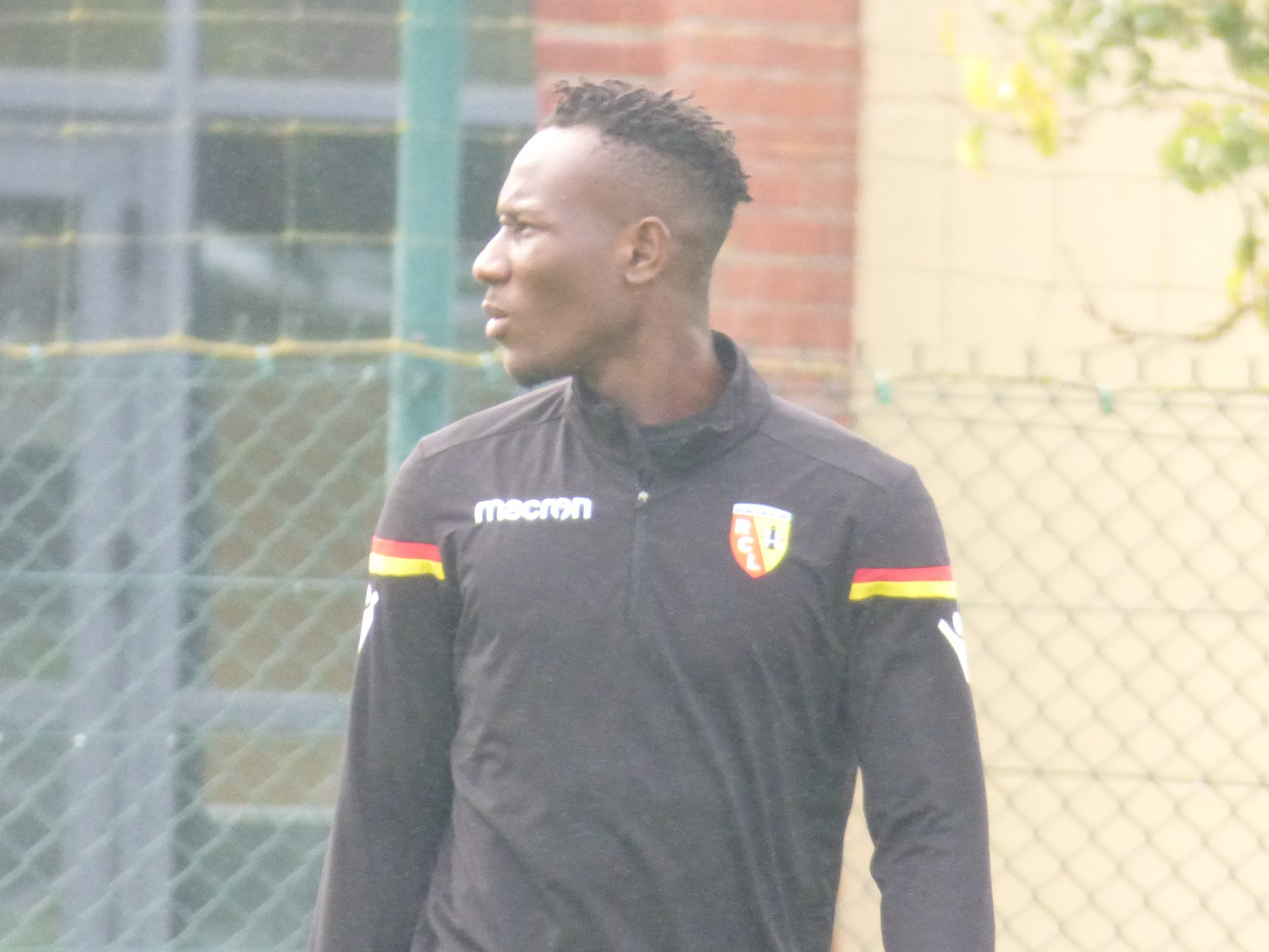 Ein Mann in einer schwarzen Jacke steht auf einem Fußballfeld, umgeben von einem Zaun und einem Baum, und sieht konzentriert während eines Trainingssitzungs.