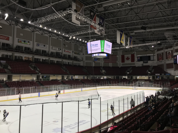 Ein Eishockeyspiel in einer großen Arena mit einem zentralen Netz, Zuschauern auf beiden Seiten und Bannern und Fahnen an den Wänden, identifiziert als die St. Louis Blues Arena.
