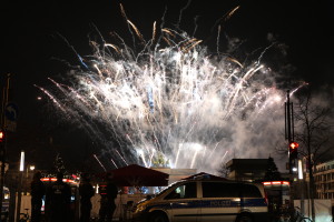 Ein Polizeiwagen steht vor einer Menschenmenge, mit beleuchteten Gebäuden und Bäumen im Hintergrund, während farbige Feuerwerke den Himmel erhellen, bei einer Silvesterfeier in Berlin.