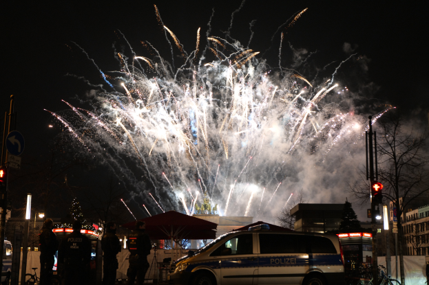 Ein Polizeiwagen steht vor einer Menschenmenge, mit beleuchteten Gebäuden und Bäumen im Hintergrund, während farbige Feuerwerke den Himmel erhellen, bei einer Silvesterfeier in Berlin.
