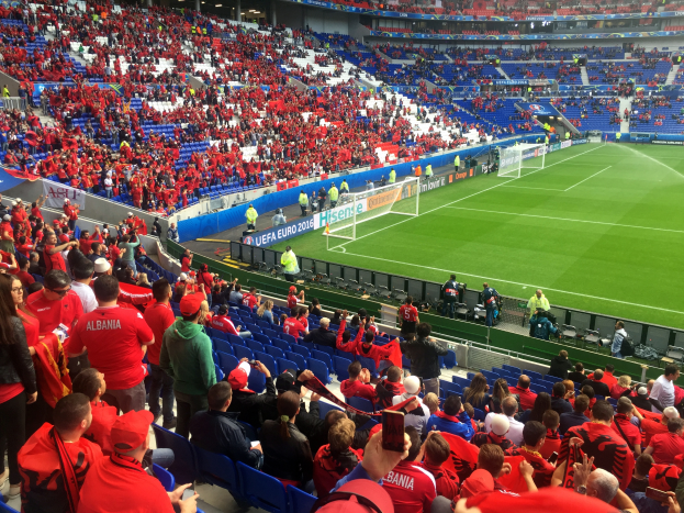Ein volles Stadion mit Zuschauern, einige sitzen und einige stehen, die ein Fußballspiel mit Torpfosten und Bannern beobachten, unter einer Decke mit Deckenleuchten.