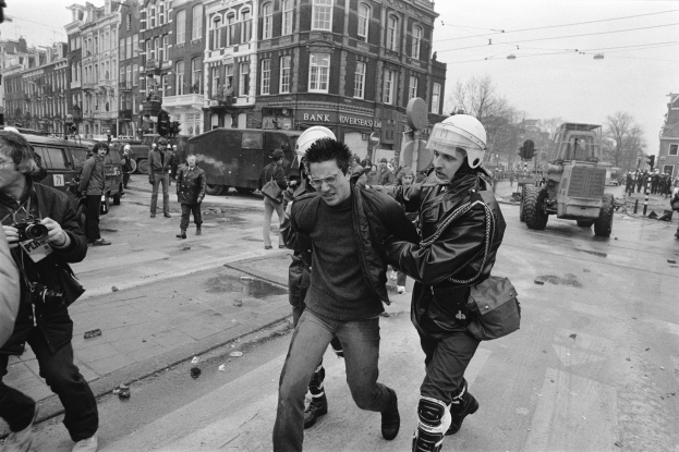 Ein Schwarz-Weiß-Bild eines Mannes, der von einem Polizeibeamten auf der Straße eskortiert wird, mit einer Gruppe von Menschen mit Kameras, Fahrzeugen, Schildern, Straßenlaternen, Stromleitungen, Bäumen, Gebäuden mit Fenstern und einer bewölkten Himmel.