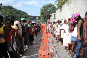 Eine lange Schlange von Menschen steht vor einem Zaun, mit einer Wand auf der rechten Seite, die auf eine Abstimmung an einer Wahllokal wartet, mit Bäumen, Gebäuden, Pfählen, Drähten und einer Wand im Hintergrund und dem Himmel oben sichtbar.