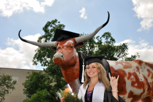 Eine Frau in einer Graduationskappe und -robe lächelt und hält einen Blumenstrauß, mit einer Skulptur, Bäumen, einem Gebäude und einem bewölkten Himmel im Hintergrund.