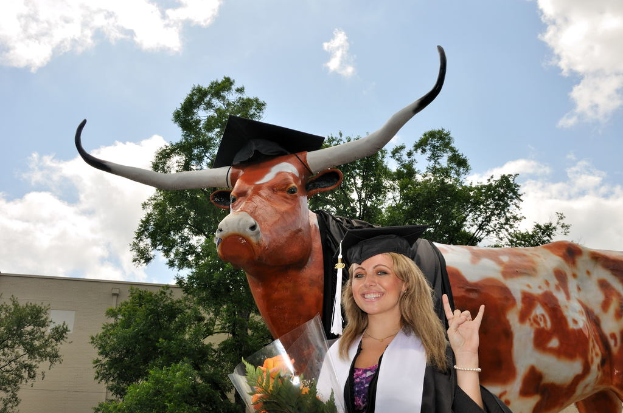 Eine Frau in einer Graduationskappe und -robe lächelt und hält einen Blumenstrauß, mit einer Skulptur, Bäumen, einem Gebäude und einem bewölkten Himmel im Hintergrund.