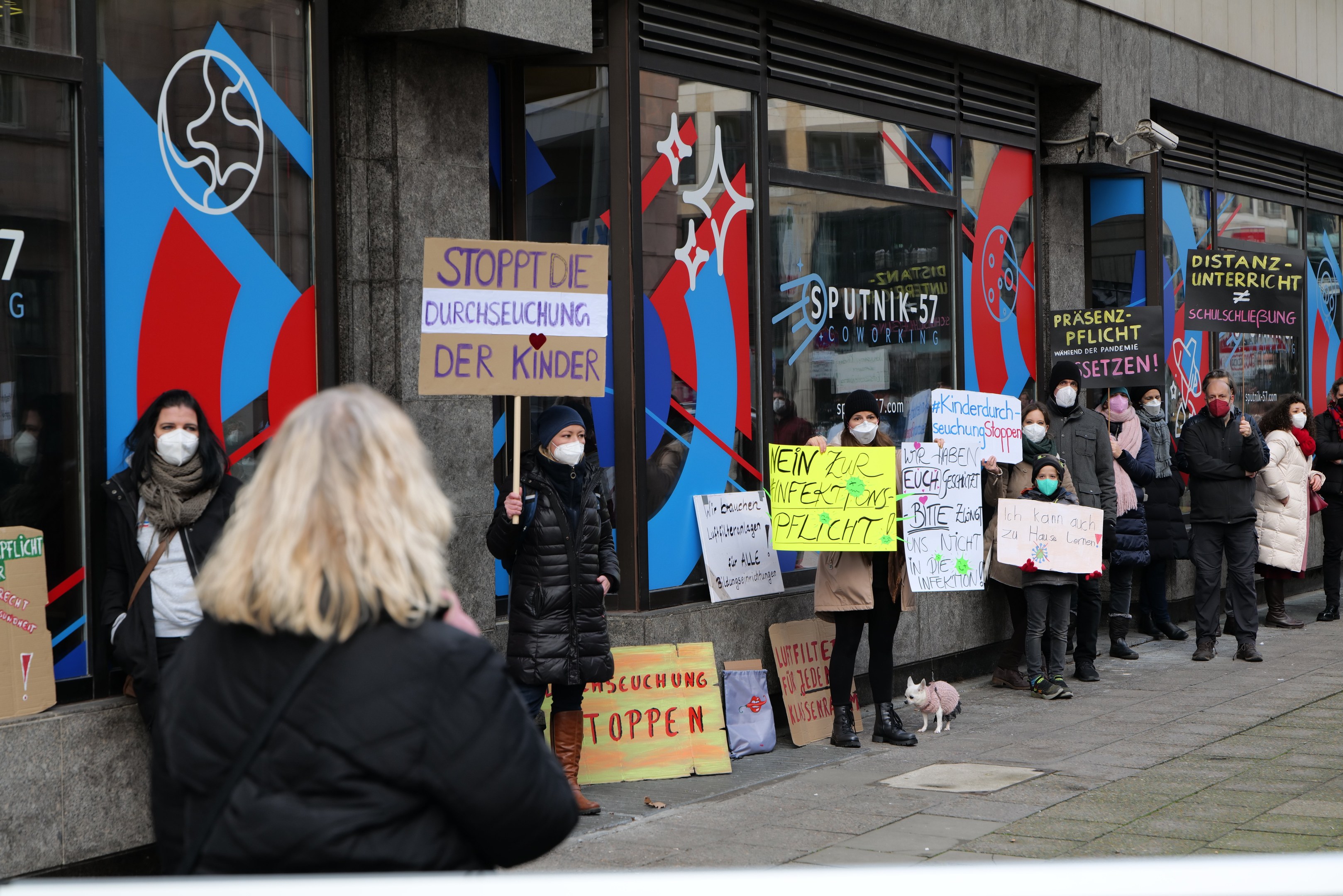Gruppe von maskierten Menschen mit Schildern und Plakaten vor einem Gebäude mit Glaswänden und -türen, mit Kameras und einem Hund, die gegen ein deutsches Maskenverbot in Schulen protestieren.