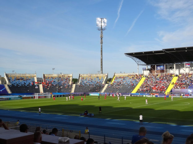 Ein Fußballspiel in einem großen, flutlichtbeleuchteten Stadion mit Zuschauern auf den Tribünen und Spielern auf dem Feld unter einem sichtbaren Himmel.