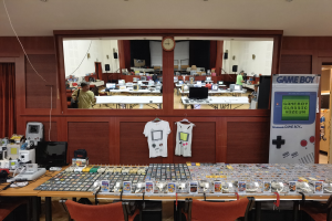 Ein Raum voller Tische und Stühle, auf denen verschiedene Gegenstände wie T-Shirts, Monitore und elektronische Geräte ausgebreitet sind, mit einer Uhr an der Wand und einem Eingang zu einem anderen Raum im Hintergrund, Teil des Nintendo Game Boy Classic Museums.