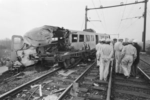Eine Schwarz-Weiß-Szene eines Zugunglücks mit Menschen in Helmen drumherum, Strommasten, Bäume, Gebäude und einen klaren Himmel im Hintergrund.