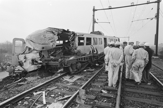 Eine Schwarz-Weiß-Szene eines Zugunglücks mit Menschen in Helmen drumherum, Strommasten, Bäume, Gebäude und einen klaren Himmel im Hintergrund.