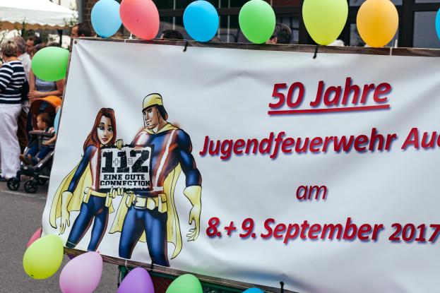 Eine Gruppe von Menschen steht auf der Straße, einige halten Kinderwagen, mit einem Banner "50 Jahre Jugendfeuerwehr August 2017" mit Text und Cartoon-Bildern, einem Zaun, Luftballons, einem Zelt, einem Gebäude mit Fenstern und Bäumen im Hintergrund.