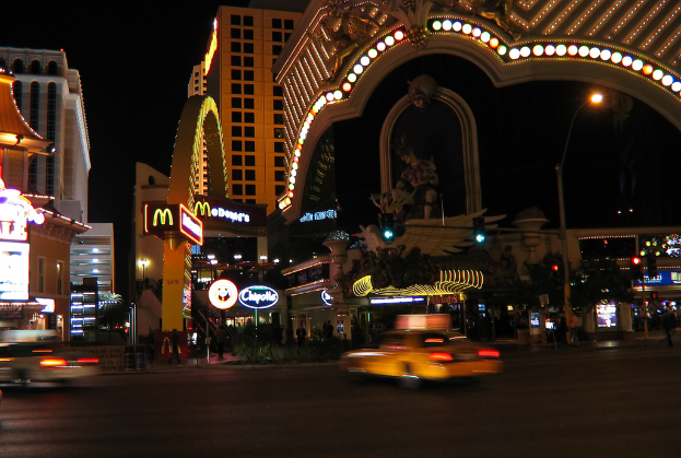 Eine nächtliche Stadtansicht mit Gebäuden, Ampeln, einer Straßenlaterne, einer Statue, Bannern und Lichtern unter einem dunklen Himmel.