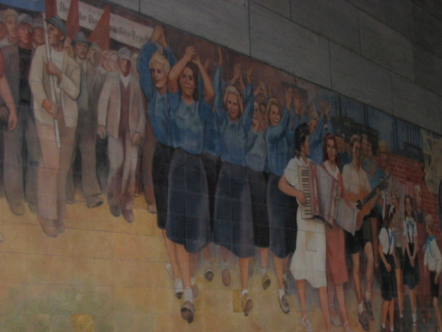 Eine Gruppe von Menschen versammelte sich, mit blauen-kleideten Mädchen in der Mitte, die klatschten, Musikern, die Instrumente spielten, Kindern, die in der rechten Ecke spielten, und Individuen, die rote Fahnen in der linken Ecke hielten.