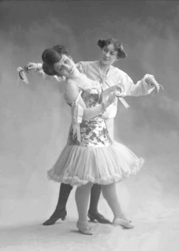 Zwei Frauen in einem Schwarz-Weiß-Foto tanzen synchron, eine hält ein Band, trägt koordinierte Kleider.