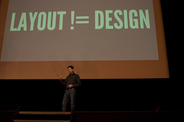 Eine Person steht auf einer Bühne und spricht, mit einem Bildschirm im Hintergrund.