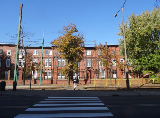 Eine Person öberquert eine Kreuzung vor einem Backsteingebäude, mit Laternenmästen, Bäumen und anderen Gegenständen drumherum und der Himmel im Hintergrund.