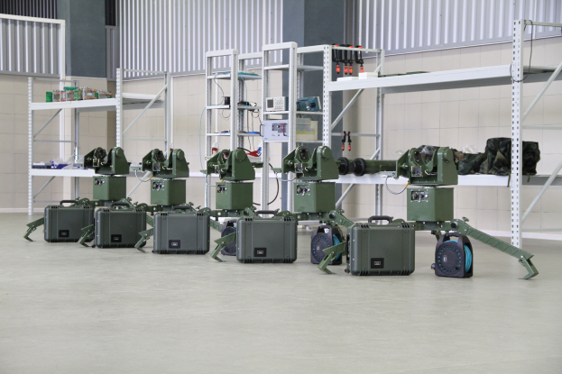 Eine Gruppe von Milit├Ąrrobotern auf einem Boden mit Koffern und Gegenst├Ąnden auf Gestellen, vor einer Wand mit einem Gel├Ąnder im Hintergrund.