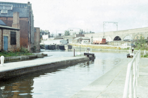 Ein altes Foto eines Kanals mit einer Brücke im Hintergrund, einem Geländer auf der rechten Seite, Gebäuden mit Fenstern und Türen auf der linken Seite und Fahrzeugen, Pfählen, Bäumen und einem klaren blauen Himmel in der Ferne.