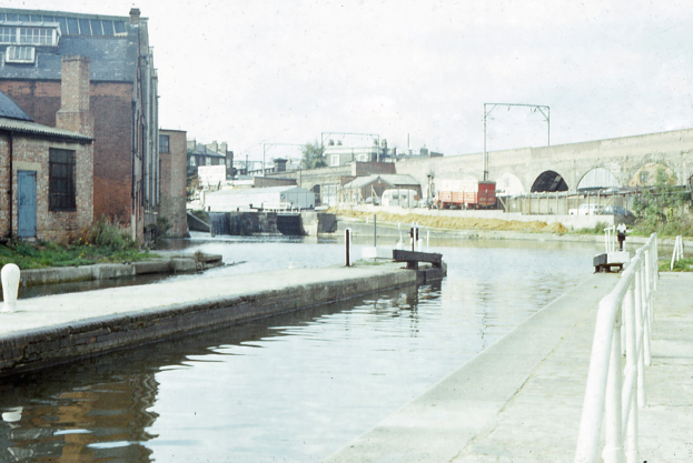Ein altes Foto eines Kanals mit einer Brücke im Hintergrund, einem Geländer auf der rechten Seite, Gebäuden mit Fenstern und Türen auf der linken Seite und Fahrzeugen, Pfählen, Bäumen und einem klaren blauen Himmel in der Ferne.