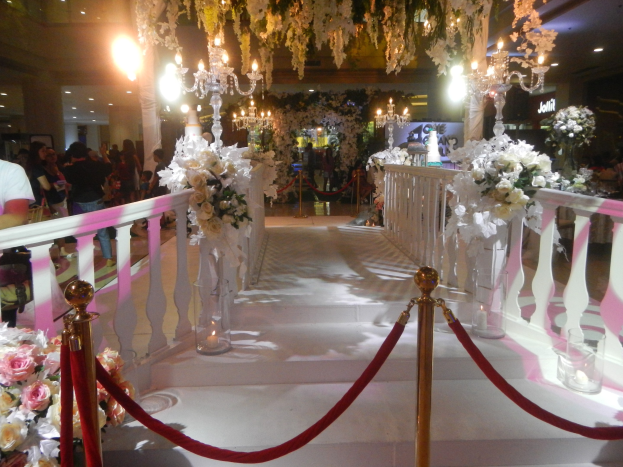 Eine Hochzeitszeremonie in einem Einkaufszentrum mit weißem Teppich, dekorativen Geländern, Blumenarrangements und Deckenbeleuchtung, bei der zahlreiche Gäste anwesend sind.