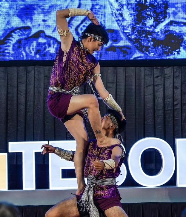 Zwei Performer in purpurfarbenen Kleidern führen einen akrobatischen Hebemanöver auf der Bühne vor einem Publikum aus, mit einem schwarzen Vorhang und einer Leinwand im Hintergrund und sitzenden Zuschauern darunter.