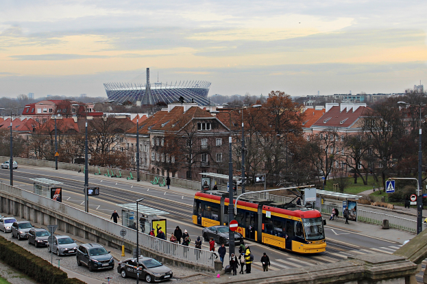 Eine gelbe und schwarze Straßenbahn fährt auf einer von Hochhäusern gesäumten Stadtstraße, umgeben von Autos, Fußgängern, Laternen, Schildern, Bäumen und Gras unter einem bewölkten Himmel.