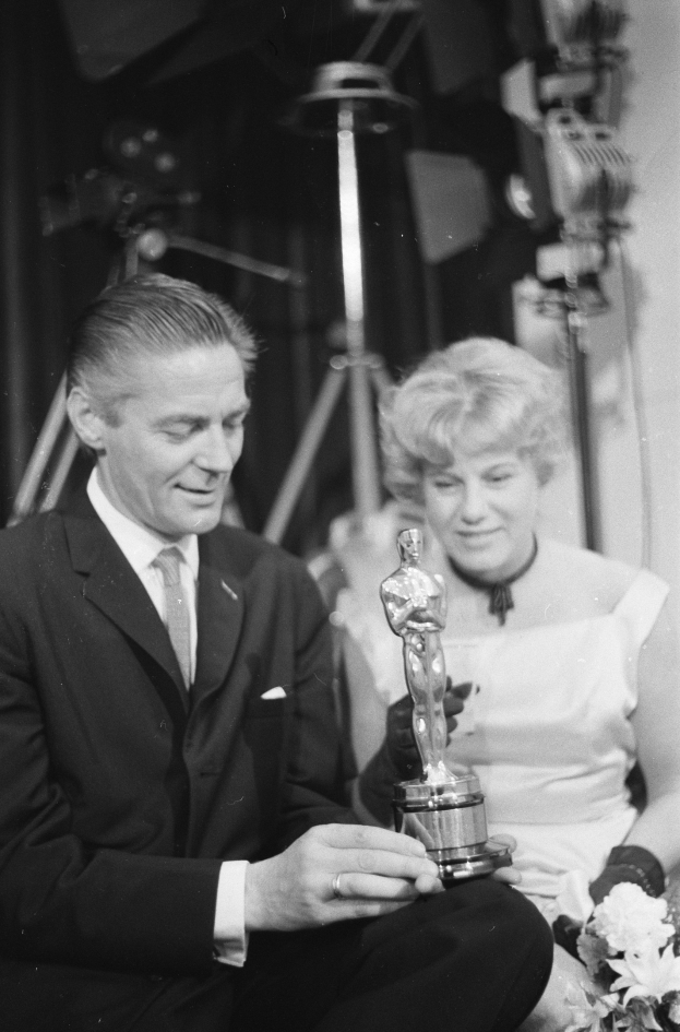 Ein Mann in einem schwarzen Blazer, weißem Hemd und Krawatte hölt eine Oscar-Statue, waehrend er neben einer Frau in Abendgarderobe sitzt, die einen Blumenstraußel hölt, mit Hintergrundbeleuchtung in einem Schwarz-Weiß-Bild.