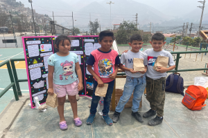 Eine Gruppe von Kindern steht auf einem Basketballfeld, jedes hält einen Papiertüte in der Hand, mit Tüten und Gegenständen rechts daneben und einer Tafel mit Text links daneben, mit Zaun, Pfosten, Straßenlaternen, Bäumen, Gebäuden, Hügeln und dem Himmel im Hintergrund.