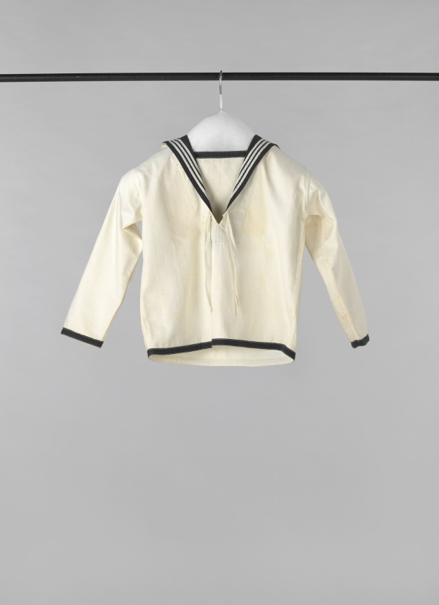 Eine weiße Matrosenjacke mit Kragen, langen Ärmeln und einem Knopfverschluss hängt an einer Wäscheleine vor einem weißen Hintergrund und besteht aus leichtem, geripptem Stoff.
