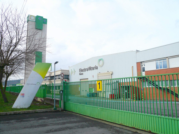 Ein großes Gebäude mit einem grünen Zaun davor, umgeben von einer Straße, Gras, Bäumen, Laternenmasten und einem Schild, unter einem bewölkten Himmel, identifiziert als Hauptsitz von Electrovictoria.