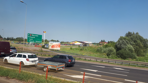 Eine Autobahn mit Autos, einem Metallzaun, Gras, Pflanzen, Bäumen, Schildern, Straßenlaternen und einem bewölktem Himmel.