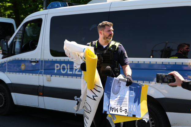 Ein Polizeibeamter in Uniform hält Papiere, während er vor einem Polizeiwagen mit Bäumen im Hintergrund steht und eine Person mit einer Kamera auf der rechten Seite zu sehen ist, was auf eine Demonstration in Berlin hindeutet.