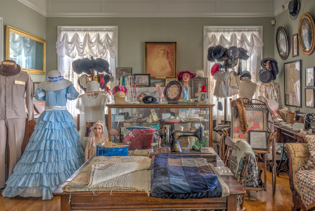 Ein Raum im Museum of Fine Arts in St. Louis, Missouri, gefüllt mit Puppen in Kleidern und Hüten, einem Tisch mit Kleidung und einer Nähmaschine, Stühlen mit Kissen, einem Schrank mit diversen Gegenständen, Fotorahmen an der Wand, Fenstern mit Vorhängen und einer Decke.