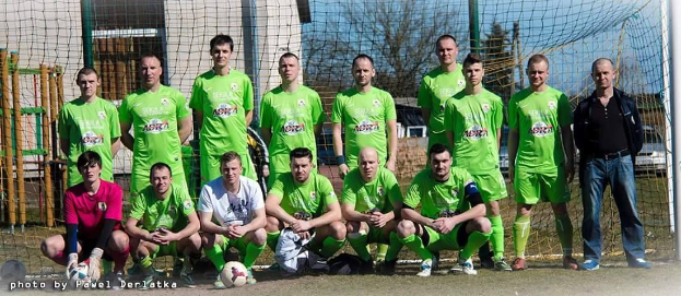 Mannergruppe in grünen T-Shirts auf einem Fußballfeld mit Ball und Tor, Bäumen und Gebäuden im Hintergrund.