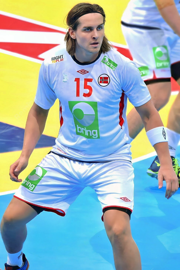 Ein Mann in weißer Uniform spielt Handball, zwei weitere Personen stehen im Hintergrund auf einem bunten Boden.