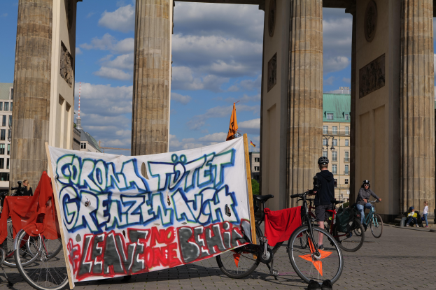 Eine Gruppe von Menschen auf Fahrrädern, die Helme tragen, fährt vor dem Brandenburger Tor in Berlin, Deutschland, mit einer Fahne und Gebäuden im Hintergrund bei einem bewölktem Himmel.