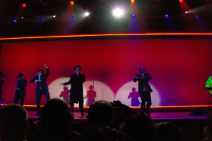 Ein Mann in einem schwarzen Kleid hält ein Mikrofon auf der Bühne, während drei Personen neben ihm tanzen, ein anderer Mann spielt rechts ein Piano, ein Kameramann filmt die Szene und Zuschauer schauen von der Seite zu, alles unter fokussierten Scheinwerfern.