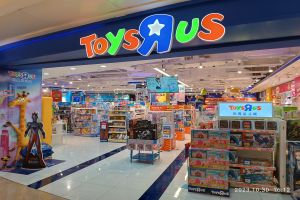 Ein Toys R Us Geschäft in einem Einkaufszentrum mit Regalen voller Spielzeug, einer Namensschild mit Text, Deckenleuchten und einem Wasserzeichen in der rechten unteren Ecke.