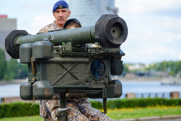 Zwei personen in militäruniformen mit grünen waffen, vor einem see, gebäuden, bäumen und einem klaren himmel.