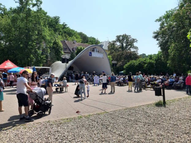 Eine Menschenmenge versammelt sich vor einem Gebäude bei der Eröffnung eines Festivals, einige sitzen auf Bänken, andere stehen, einschließlich eines Kinderwagens in der foreground; Bäume, Gebäude und ein klarer blauer Himmel sind im Hintergrund zu sehen, zusammen mit Zelten auf der linken Seite und einem Müllcontainer auf der rechten Seite.