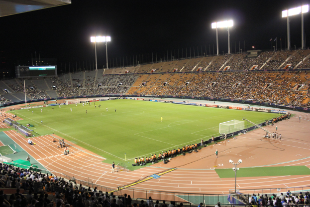 Ein großes Stadion voller Zuschauer bei einem Fußballspiel unter Flutlicht in der Nacht, mit einem Anzeigeboard im Hintergrund.