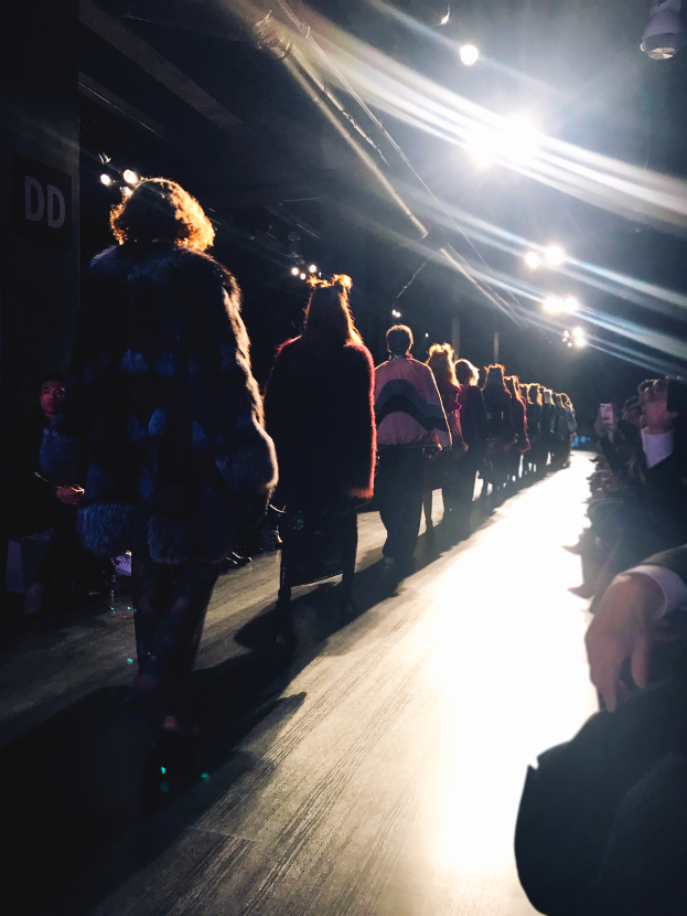 Eine Gruppe von Menschen läuft auf einem hell erleuchteten Laufsteg bei einer Modenschau, mit Zuschauern auf beiden Seiten.